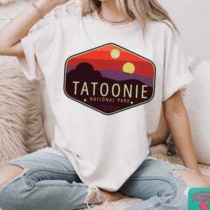 May include: Cream-colored t-shirt featuring a graphic of a sunset over a desert landscape with the text "TATOONIE NATIONAL PARK." The design includes two suns and the phrase "Twice the Sun, Twice the Fun." The shirt is made of a soft material.