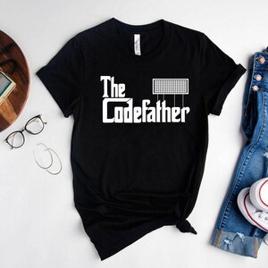 May include: A black short-sleeved t-shirt featuring white screen-printed text that reads The Codefather, with a small grid-like graphic positioned above the word Codefather. The shirt has a crew neckline and a standard fit design.
