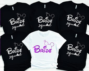 May include: A collection of five short-sleeved t-shirts. Four are black with white text reading Bride squad and a mouse ear silhouette with a diamond ring. One is white with purple text reading Bride and a mouse ear silhouette with a diamond ring.