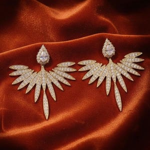May include: A pair of gold-toned earrings with a leaf-like design, encrusted with small, sparkling crystals. Each earring features a teardrop-shaped crystal at the top. The earrings are displayed on a rust-colored velvet surface.