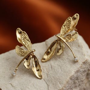 Op de afbeelding: Een paar goudkleurige oorringen in de vorm van een libelle. Elke oorring heeft gedetailleerde vleugels en een delicate body, met een kleine, heldere edelsteen aan het einde van de body. De oorbellen worden gepresenteerd op een lichtgekleurd, getextureerd oppervlak.