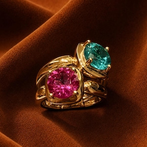 May include: Two gold rings stacked together, one featuring a round, vibrant pink gemstone and the other featuring a round, bright teal gemstone. Both rings have polished gold bands and four-prong settings that secure the faceted stones in place.