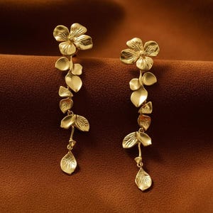 May include: A pair of gold-colored drop earrings. Each earring features a floral design at the top, with a series of leaves and a teardrop-shaped pendant. The earrings are made of a shiny, metallic material.