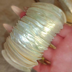 May include: A pearlescent bracelet composed of numerous shell-like segments. The bracelet has a natural, iridescent sheen, reflecting light with a subtle, shimmering effect. The design suggests a coastal or beach-inspired theme.