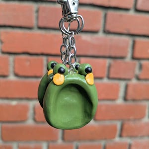May include: A green frog-shaped keychain with black eyes and orange accents. The frog is attached to a silver chain and clasp. The background is a red brick wall.