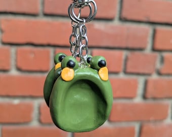 May include: A green frog-shaped keychain with black eyes and orange accents. The frog is attached to a silver chain and clasp. The background is a red brick wall.