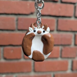 May include: A handmade keychain featuring a round, white and brown hedgehog-like creature with small antlers. The charm is attached to a silver-colored chain and clasp. The background is a red brick wall.