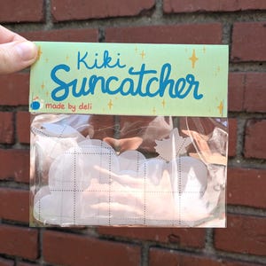 Puede incluir: Un paquete de atrapasoles Kiki Suncatcher. El embalaje es de color verde claro con las palabras "Kiki Suncatcher" en escritura azul. Los atrapasoles tienen forma de nube y son visibles a través del embalaje de plástico transparente. El fondo es una pared de ladrillos rojos.