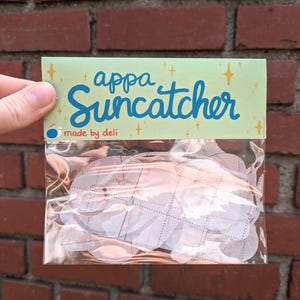 Puede incluir: Una bolsa de plástico transparente que contiene múltiples atrapasoles appa. El paquete del atrapasoles tiene un encabezado verde claro con las palabras "appa Suncatcher" en escritura azul. Las palabras "made by deli" están impresas debajo. Los atrapasoles son blancos y tienen un patrón acolchado.