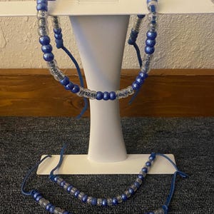 Pode incluir: Um conjunto de colar e pulseira com contas transparentes e azuis. As contas são enfiadas num cordão azul. O colar é exibido num suporte branco. O fundo inclui elementos castanhos e cinzentos.