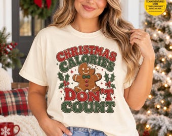 May include: A cream-coloured short-sleeve t-shirt featuring a graphic of a gingerbread man with a bite taken out of its head. The text reads CHRISTMAS CALORIES DON'T COUNT in red and green, surrounded by holly leaves and berries.