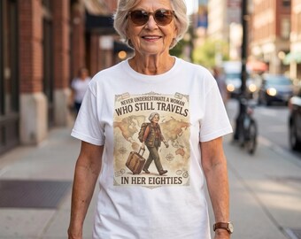 Op de afbeelding: Een wit T-shirt met korte mouwen en een grafische print van een reiziger met een koffer en rugzak op een wereldkaartachtergrond. De tekst luidt: NEVER UNDERESTIMATE A WOMAN WHO STILL TRAVELS IN HER EIGHTIES, omlijst door decoratieve randen.