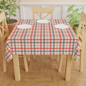 May include: A dining table set with a red, white, gray, and tan plaid tablecloth. Three white plates are on the table. The table is surrounded by four light wood chairs. Potted plants are in the background.