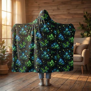 Puede incluir: Una manta con capucha negra con un patrón repetido de hongos verdes y azules brillantes, hojas y estrellas. La manta está sobre una persona, con la capucha puesta. El fondo es un interior acogedor con pisos de madera y una ventana.