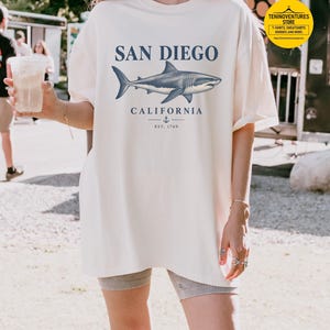 Woman wearing an ivory Comfort Colors 1717 T-shirt featuring a vintage navy shark illustration, San Diego California lettering, and a small anchor on a white background.