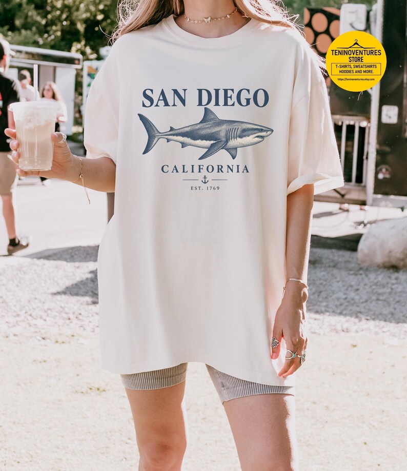 Woman wearing an ivory Comfort Colors 1717 T-shirt featuring a vintage navy shark illustration, San Diego California lettering, and a small anchor on a white background.