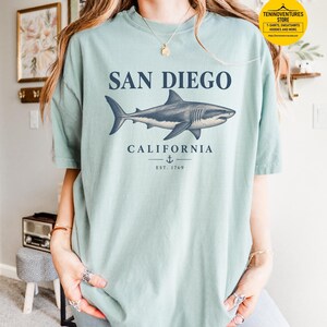 Woman wearing a bay green Comfort Colors 1717 T-shirt featuring a vintage navy shark illustration, San Diego California lettering, and a small anchor on a white background.