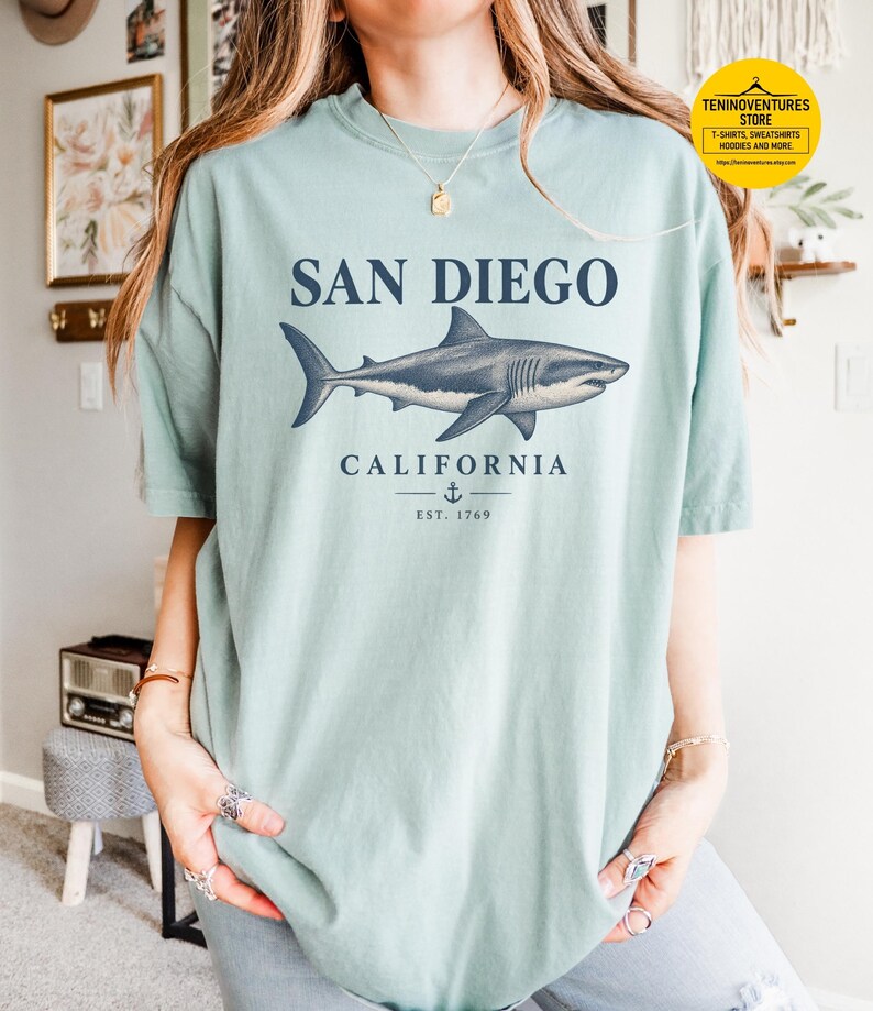 Woman wearing a bay green Comfort Colors 1717 T-shirt featuring a vintage navy shark illustration, San Diego California lettering, and a small anchor on a white background.