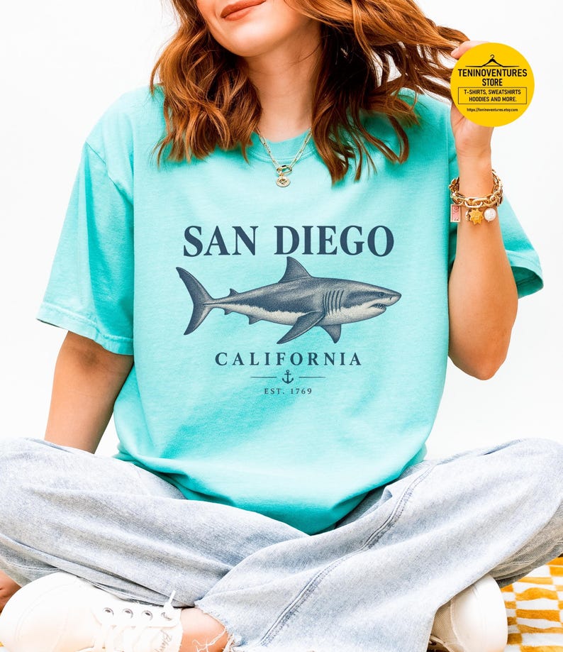 Woman wearing a chalky mint Comfort Colors 1717 T-shirt featuring a vintage navy shark illustration, San Diego California lettering, and a small anchor on a white background.