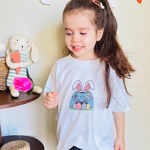 May include: A white t-shirt featuring a blue video game controller with bunny ears and Easter eggs. The shirt is worn by a young child. A decorative garland with carrots and bunnies hangs in the background.