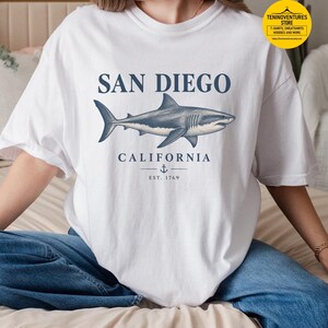 Woman wearing a white Comfort Colors 1717 T-shirt featuring a vintage navy shark illustration, San Diego California lettering, and a small anchor on a white background.