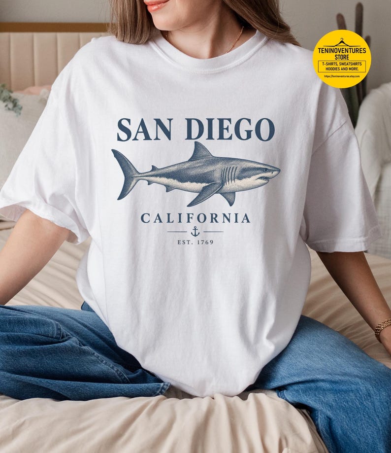 Woman wearing a white Comfort Colors 1717 T-shirt featuring a vintage navy shark illustration, San Diego California lettering, and a small anchor on a white background.