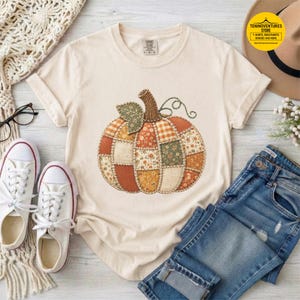 May include: A cream-coloured short-sleeve t-shirt featuring a large central graphic of a pumpkin composed of various patchwork fabric patterns in shades of orange, green, and brown. The shirt has a crew neckline and a tag at the inner collar.