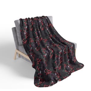 May include: A dark gray throw blanket draped over a gray armchair. The blanket features a repeating pattern of black roses, red berries, and silver swords with red ribbons. The blanket is made of a soft material.