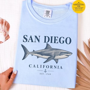 Folded flat lay of a hydrangea blue Comfort Colors 1717 T-shirt featuring a vintage navy shark illustration, San Diego California lettering, and a small anchor on a white background.