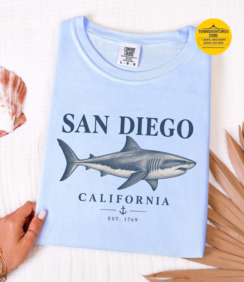 Folded flat lay of a hydrangea blue Comfort Colors 1717 T-shirt featuring a vintage navy shark illustration, San Diego California lettering, and a small anchor on a white background.