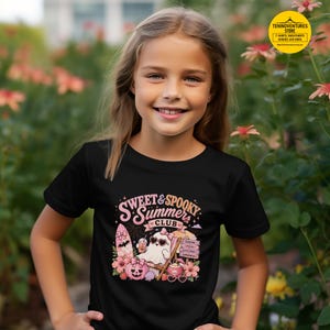 May include: Black t-shirt with the text "Sweet & Spooky Summer Club" and a graphic of a ghost wearing sunglasses, a pumpkin, and flowers. The shirt is worn by a young girl smiling at the camera.