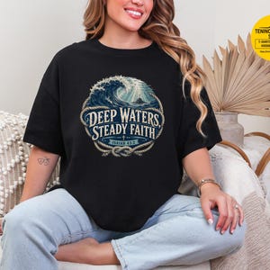 May include: A black short-sleeve t-shirt featuring a circular graphic of a large ocean wave and a cross under light rays. The design is framed by a rope border and includes the text DEEP WATERS, STEADY FAITH and ISAIAH 43:2 in white lettering.