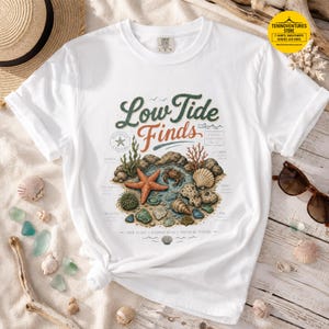 May include: A white short-sleeve t-shirt featuring a graphic print with the text Low Tide Finds. The illustration depicts a coastal scene with a starfish, crab, various seashells, sea glass, and coral, labeled with small descriptive text.