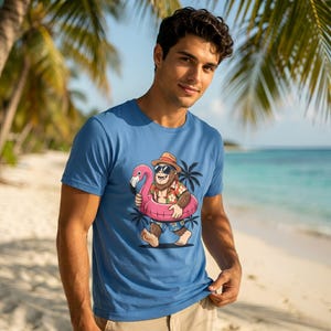 Puede incluir: Una camiseta azul con un personaje de dibujos animados de Bigfoot con gafas de sol, una camisa hawaiana y pantalones cortos, flotando en un flotador de piscina de flamenco rosa. El Bigfoot también lleva un sombrero. El fondo muestra palmeras, una playa y el océano.