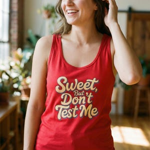 Könnte beinhalten: Ein rotes Tanktop mit dem Text "Sweet, But Don't Test Me" in einer Retro-Schriftart. Der Text ist cremefarben mit einer dunkelblauen Umrandung. Das Tanktop hat einen Rundhalsausschnitt und ist aus weichem Material.