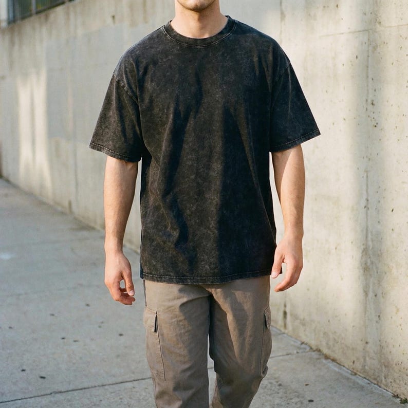 A person wearing a short-sleeve, black acid wash oversized t-shirt and grey cargo pants, standing outdoors against a concrete wall.