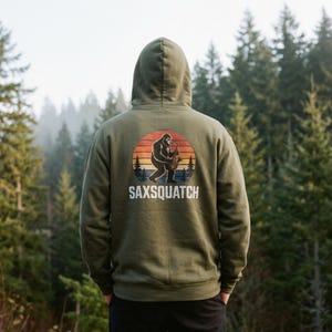 May include: Olive green hoodie with a graphic of a Bigfoot playing a saxophone against a sunset backdrop. The text "SAXSQAUTCH" is below the graphic. The hoodie is made of a soft material and has a hood.