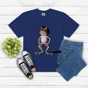 May include: Navy blue t-shirt featuring a cartoon frog with a bob haircut. The frog is green with a pink belly and black spots. The image also includes blue jeans and black canvas sneakers.