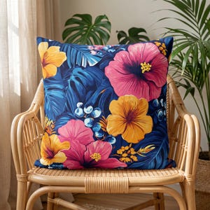 Könnte beinhalten: Ein quadratisches Zierkissen mit einem lebendigen tropischen Muster aus großen rosa und gelben Hibiskusblüten vor einem tief dunkelblauen Hintergrund aus üppigen Palmenblättern und kleinen blauen Beeren in einem auffälligen botanischen Design.