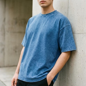 A person leaning against a concrete wall, wearing a short-sleeve dark blue acid wash oversized t-shirt with hands in pockets.