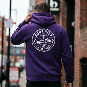 May include: A purple hoodie with a white circular graphic on the back. The graphic reads "Surf City Santa Cruz California" with an anchor symbol. The hoodie has a drawstring hood and long sleeves. The person is wearing black pants.