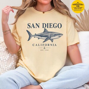 Woman wearing a butter Comfort Colors 1717 T-shirt featuring a vintage navy shark illustration, San Diego California lettering, and a small anchor on a white background.