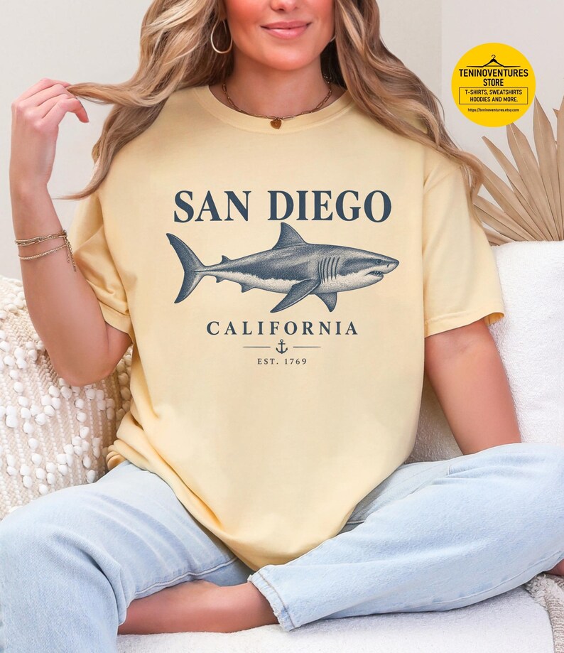 Woman wearing a butter Comfort Colors 1717 T-shirt featuring a vintage navy shark illustration, San Diego California lettering, and a small anchor on a white background.
