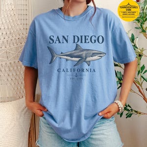 Woman wearing a washed denim Comfort Colors 1717 T-shirt featuring a vintage navy shark illustration, San Diego California lettering, and a small anchor on a white background.