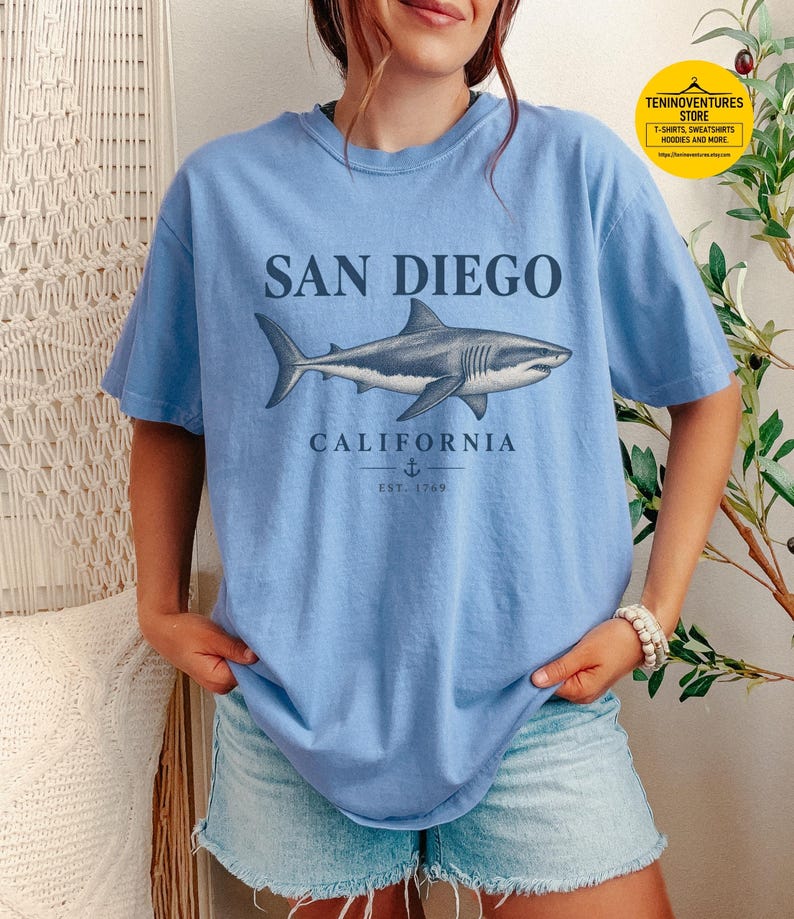 Woman wearing a washed denim Comfort Colors 1717 T-shirt featuring a vintage navy shark illustration, San Diego California lettering, and a small anchor on a white background.