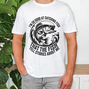 Peut inclure: T-shirt blanc avec un motif noir et blanc d'un poisson sautant hors de l'eau. Le texte sur le t-shirt se lit comme suit : "I'M SO GOOD AT CATCHING FISH THAT THE FISH TELL STORIES ABOUT ME."