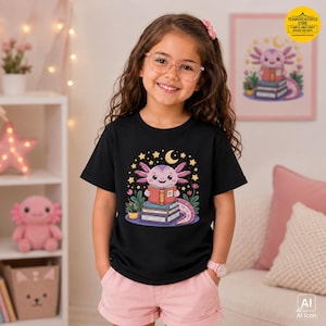 May include: A black short-sleeve t-shirt featuring a graphic of a pink axolotl reading a book while sitting on a stack of books, surrounded by yellow stars, a crescent moon, and small green plants. The design is centered on the chest.