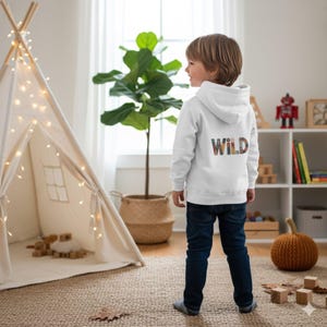 Puede incluir: Una sudadera con capucha blanca con la palabra "WILD" impresa en la espalda en una fuente colorida y estampada. La sudadera es usada por un niño con jeans azules. El fondo incluye un tipi, una planta y una calabaza.