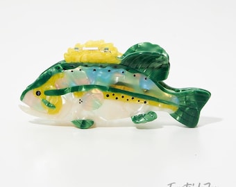 May include: A decorative fish figurine with a pearlescent body, accented with yellow, green, and blue details. The fish has a textured appearance and a yellow crest. The tail and fins are a vibrant green. The figurine is set against a white background.
