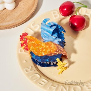 May include: A decorative rooster figurine with a vibrant color scheme of orange, blue, and yellow. The rooster is perched on a cream-colored plate, accompanied by two red cherries. The figurine has a glossy finish and detailed design.